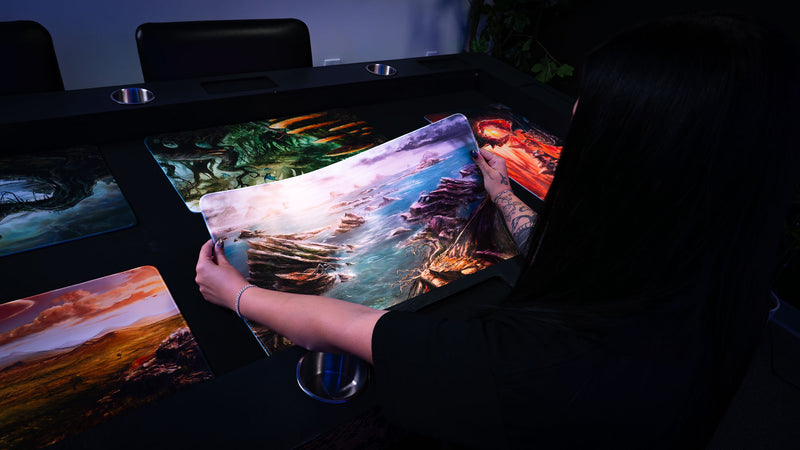 Person holding the Desolate Island playmat to demonstrate its flexible premium surface, featuring a dramatic coastal landscape with rocky sea cliffs, scattered islands, blue ocean waters, and a rugged fantasy shoreline.