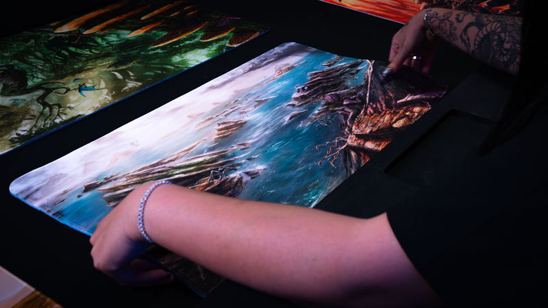 Person positioning the Desolate Island playmat on a gaming table, featuring a rugged coastal landscape with rocky cliffs, scattered islands, crashing blue waters, and a windswept fantasy shoreline.