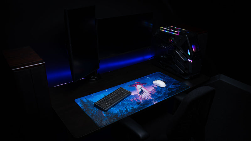 Angled view of the Dino Abduction Rust deskmat on a gaming desk, highlighting the colorful UFO artwork with keyboard and mouse in a complete desktop setup.