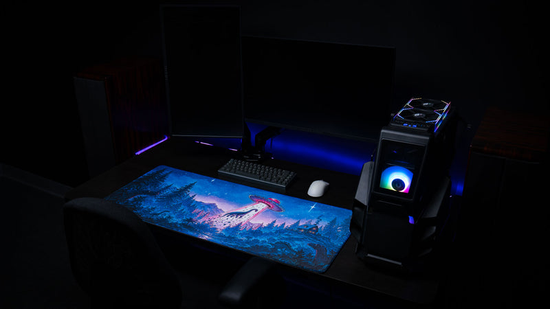 Dino Abduction Rust deskmat displayed on a modern gaming desk with dual monitors, showcasing the vibrant UFO and dinosaur artwork in a complete desktop setup.