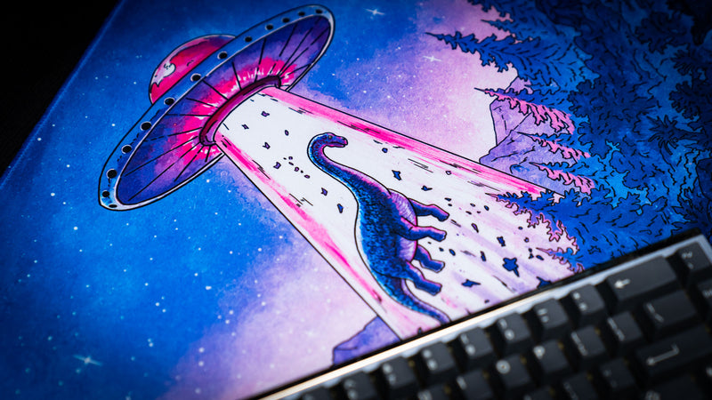 Close-up of the Dino Abduction Rust deskmat featuring a glowing UFO beaming up a dinosaur against a vibrant starry night sky.
