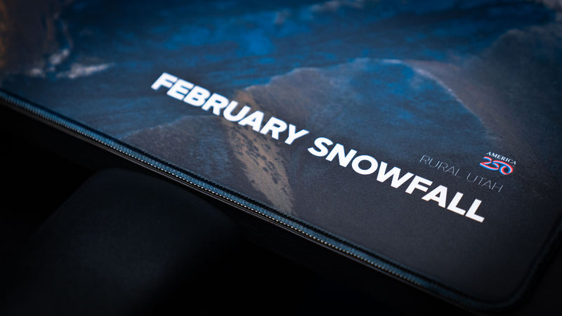 Close-up of the February Snowfall deskmat title and America 250 logo printed on the premium stitched-edge gaming deskmat.