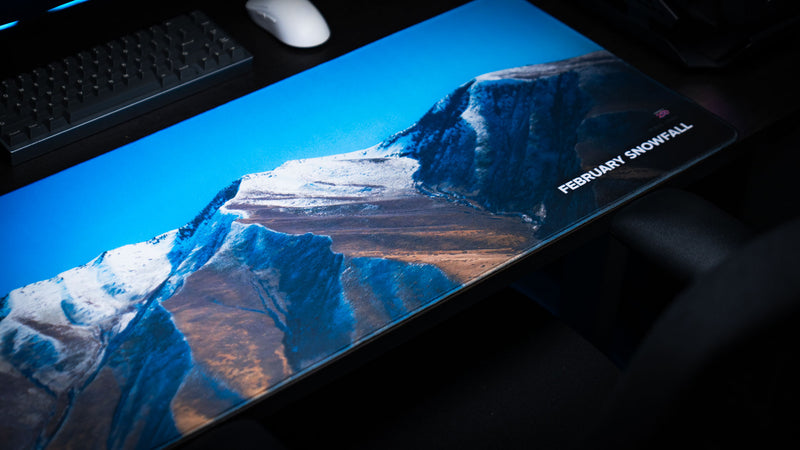 February Snowfall deskmat featuring snow-covered Utah mountains on a gaming desk with a blue sky panorama and premium stitched edges.