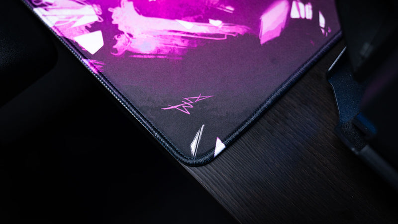 Close-up of the Forbidden Spoils gaming deskmat showing the artist signature, reinforced stitched edge, and premium cloth surface with vivid purple artwork.