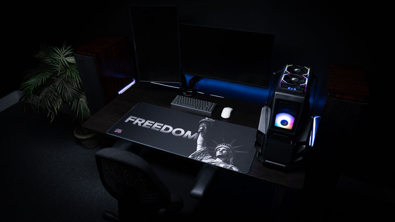 Wide desk setup featuring the Freedom deskmat with monochrome Statue of Liberty artwork and bold "FREEDOM" text, displayed beneath a mechanical keyboard and gaming PC.