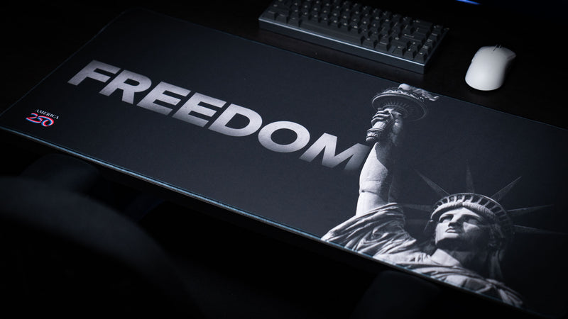 Angled overhead view of the Freedom deskmat featuring bold "FREEDOM" lettering, monochrome Statue of Liberty artwork, America 250 logo, and a premium extended gaming mouse pad setup.