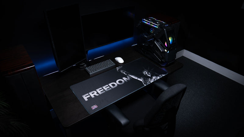 Wide gaming desk setup featuring the Freedom deskmat with bold "FREEDOM" lettering, monochrome Statue of Liberty artwork, America 250 logo, mechanical keyboard, and gaming PC.