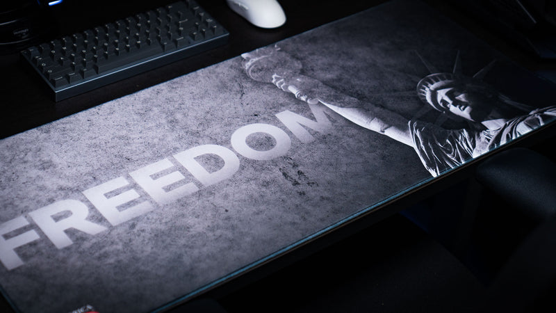Angled close-up of the Freedom deskmat showcasing bold "FREEDOM" lettering over a distressed gray background with the Statue of Liberty and stitched premium edges.