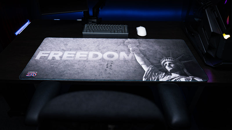 Full desk view of the Freedom deskmat featuring bold "FREEDOM" typography, a distressed grayscale Statue of Liberty design, the America 250 logo, and premium stitched edges.