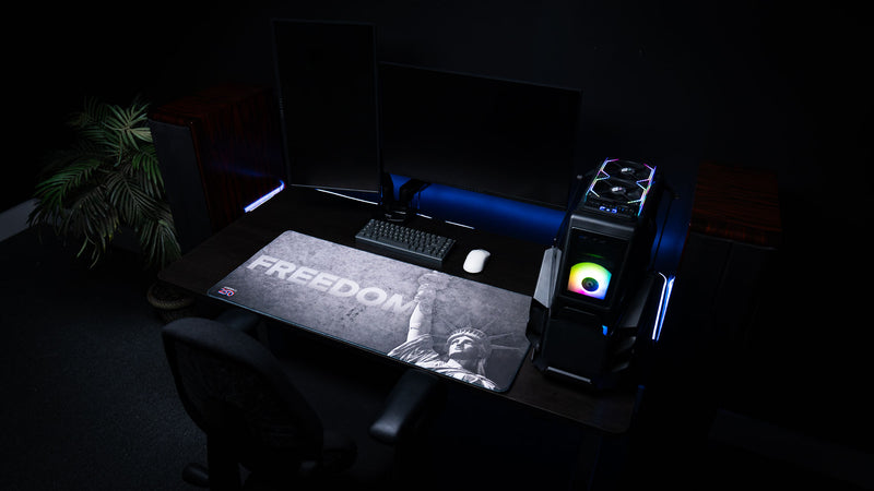 Wide-angle gaming setup featuring the Freedom deskmat with distressed grayscale artwork, bold "FREEDOM" typography, the Statue of Liberty, America 250 logo, and premium stitched edges.