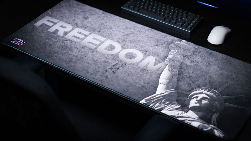 Perspective view of the Freedom deskmat featuring bold "FREEDOM" typography, distressed grayscale artwork, the Statue of Liberty, America 250 logo, and premium stitched edges.