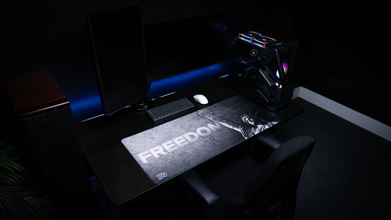 Wide gaming setup featuring the Freedom deskmat with distressed grayscale artwork, bold "FREEDOM" typography, the Statue of Liberty, America 250 logo, and premium stitched edges.