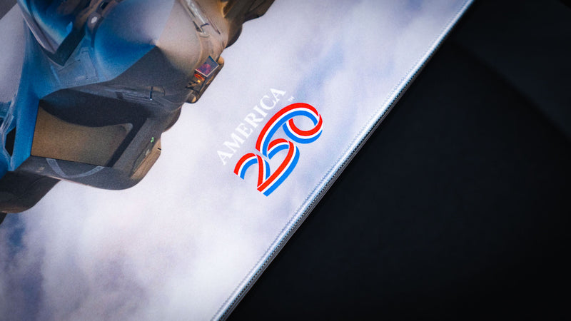 Close-up of the Freedom in the Sky America 250 deskmat showing the America 250 logo, light cloud artwork, and fighter jet detail.