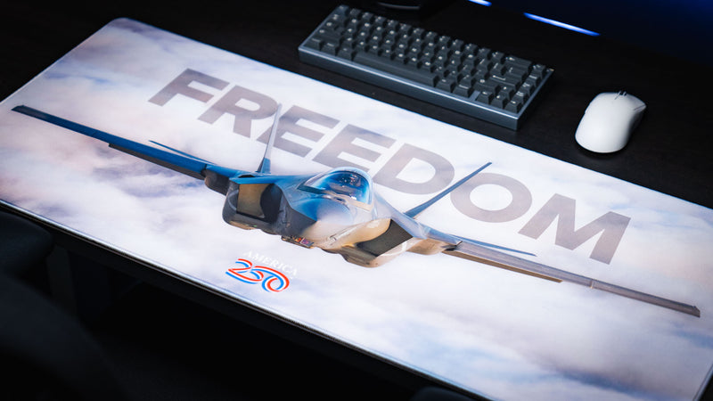 Angled view of the Freedom in the Sky America 250 deskmat featuring a fighter jet, cloudy sky design, keyboard, and white mouse.