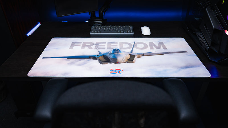 Centered workstation view of the Freedom in the Sky America 250 deskmat featuring a fighter jet, cloudy sky artwork, keyboard, mouse, and PC setup.