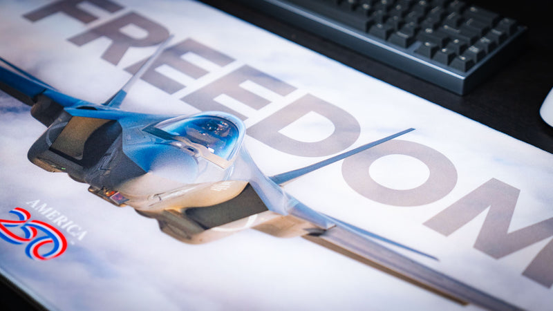 Close-up of the Freedom in the Sky America 250 deskmat featuring bold FREEDOM lettering, fighter jet artwork, and soft cloud print detail.