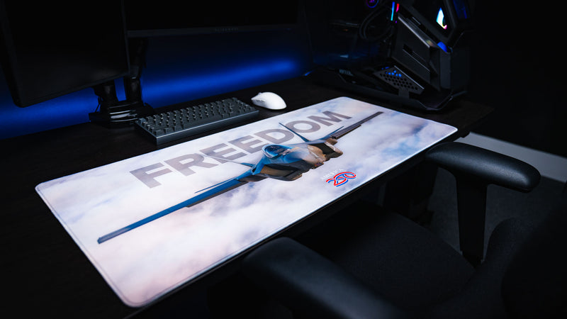 Side profile view of the Freedom in the Sky America 250 deskmat featuring a fighter jet, cloudy sky artwork, keyboard, and mouse.