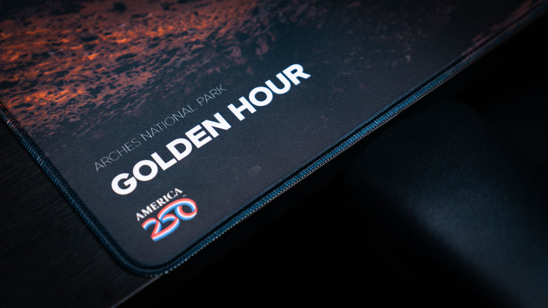 Golden Hour deskmat corner detail featuring the printed title, Arches National Park text, America 250 logo, and premium stitched border.
