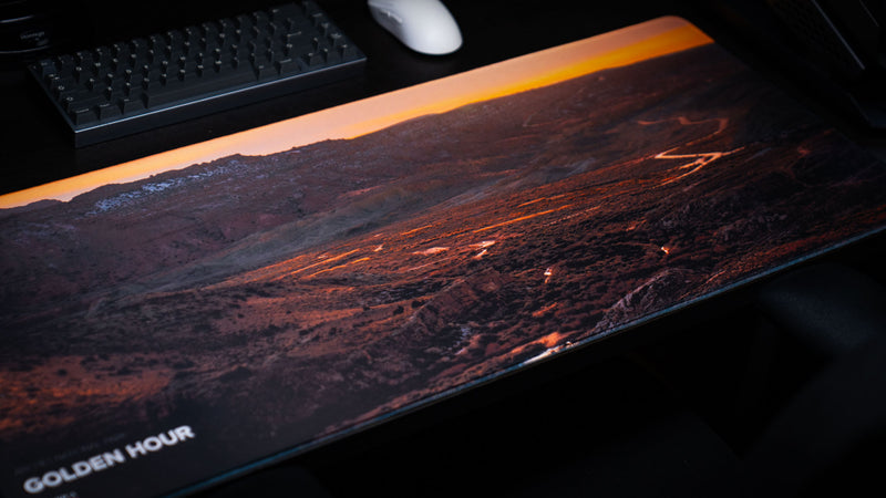 Close-up of the Golden Hour America 250 deskmat showing glowing desert valley roads, rugged Arches National Park terrain, and stitched edge detail