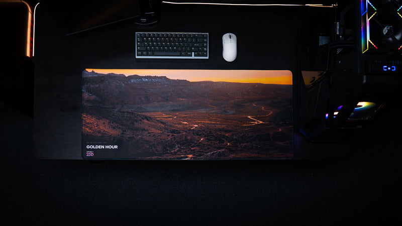 Top-down gaming desk display of the Golden Hour America 250 deskmat with sunset canyon artwork, title branding, keyboard, mouse, and PC setup.