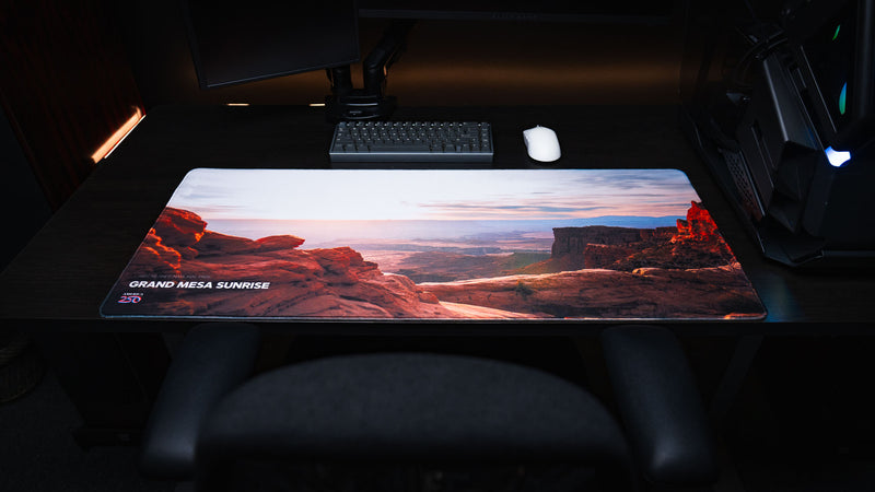 Front workstation view of the Grand Mesa Sunrise America 250 deskmat featuring Canyonlands sunrise scenery, red rock cliffs, keyboard, and mouse.