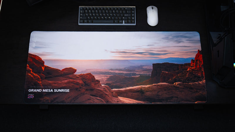 Full desktop view of the Grand Mesa Sunrise America 250 deskmat featuring Canyonlands sunrise colors, red rock formations, keyboard, and white mouse.