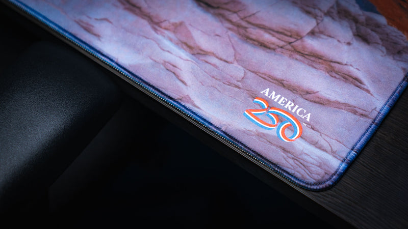 Close-up of the Grand Mesa Sunrise deskmat corner featuring the America 250 logo, premium stitched edges, and vibrant Mesa Arch sunrise photography from Canyonlands National Park.