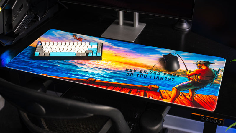 How Do You F@#K Do You Fish deskmat by Posty displayed beneath a mechanical keyboard and gaming mouse, featuring a colorful sunset ocean scene and fisherman artwork.