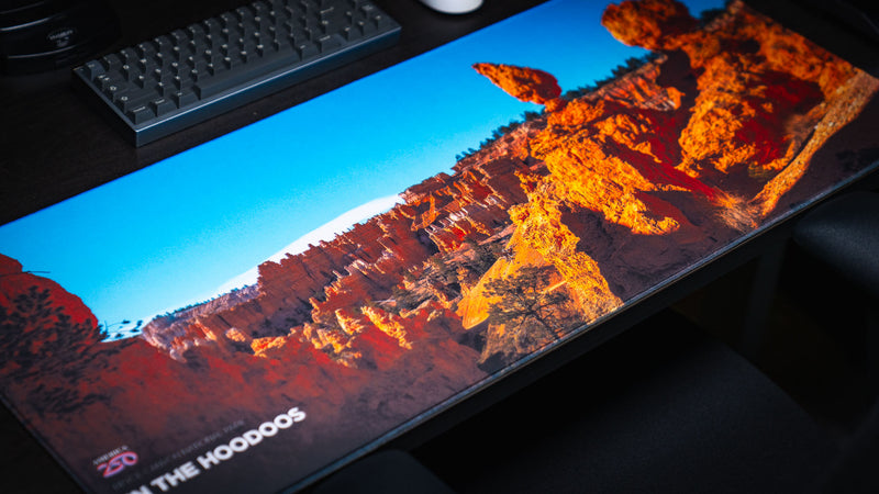 Angled view of the In the Hoodoos Bryce Canyon National Park deskmat showcasing vibrant orange hoodoo formations, clear blue sky, stitched edges, and a premium gaming desk setup.