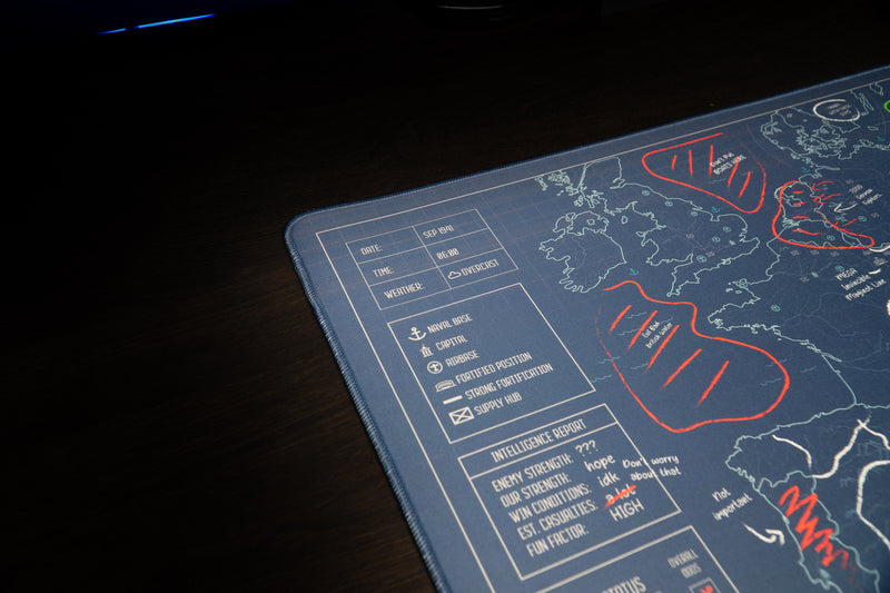 Close-up of the Jaush deskmat's stitched corner, map legend, and humorous intelligence report beside western Europe.