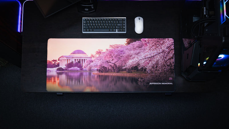 Top-down view of the America 250 Jefferson Memorial deskmat featuring the Jefferson Memorial and blooming cherry blossoms reflected in the Tidal Basin on a premium gaming desk with a mechanical keyboard and white gaming mouse.