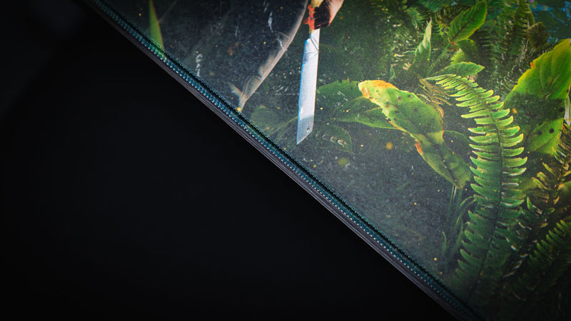 Close-up of the Jungle Exploration deskmat showcasing the premium stitched edge, detailed jungle foliage, and Rust survivor's machete.