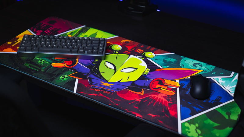 Angled view of the Lure of the Night gaming deskmat featuring a colorful comic-style moth character with keyboard and mouse on a gaming desk.