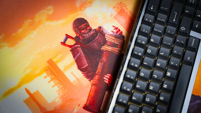 Close-up of the main scene Rust deskmat highlighting a rocket launcher raider preparing to fire during a base raid, showcasing the vivid artwork and premium stitched-edge gaming mouse pad surface beside a mechanical keyboard.