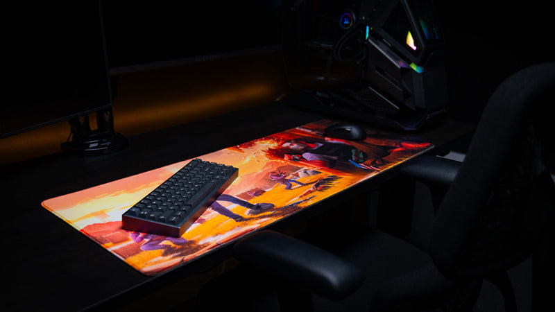 Low side-angle view of the Main scene Rust deskmat featuring a high-intensity raid scene with players exchanging gunfire and explosions, displayed as a premium extended gaming mouse pad with stitched edges beneath a mechanical keyboard and mouse.