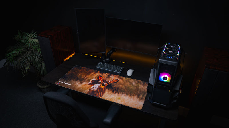 Premium Mule Deer Buck deskmat displayed on a modern gaming setup with dual monitors, mechanical keyboard, RGB PC, and a detailed wildlife photograph from Bryce Canyon National Park.