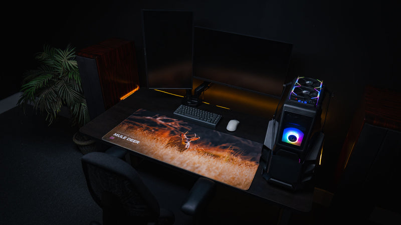 Wide desktop view of the Mule Deer America 250 deskmat featuring a mule deer in golden Zion grass with keyboard, mouse, and RGB PC setup.