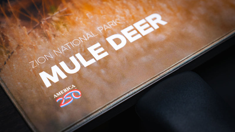 Close-up of the Mule Deer America 250 deskmat showing Zion National Park title text, America 250 logo, warm grass artwork, and stitched edge.