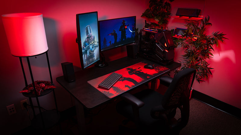 Gaming room setup featuring the Nighttime Patrol Rust deskmat with red ambient lighting, dual monitors, gaming PC, greenery, and silhouetted scientists patrolling beneath a crimson night sky.