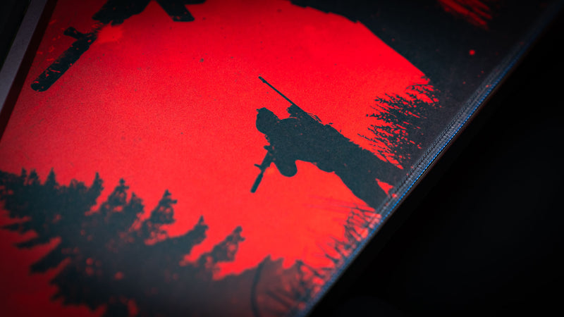 Close-up detail of the Nighttime Patrol Rust deskmat featuring a silhouetted sniper standing watch against a vivid red night sky, highlighting the textured surface, sharp print quality, and stitched edge construction.