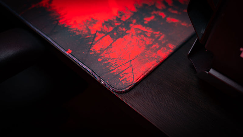 Macro close-up of the Nighttime Patrol Rust deskmat showcasing premium stitched edges, durable construction, and the dark forest silhouette artwork against a striking red background.