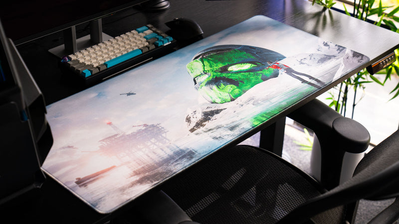 Angled view of the Oilrats Frozen Paradise deskmat featuring a massive glowing green skull emerging from a frozen landscape, with an offshore rig, helicopter, icy reflections, and a lone survivor standing in the snow.