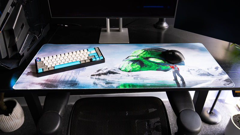 Oilrats Frozen Paradise deskmat displayed on a gaming workstation with keyboard and mouse, showcasing the frozen landscape, glowing green skull monument, and reflective ice scene.