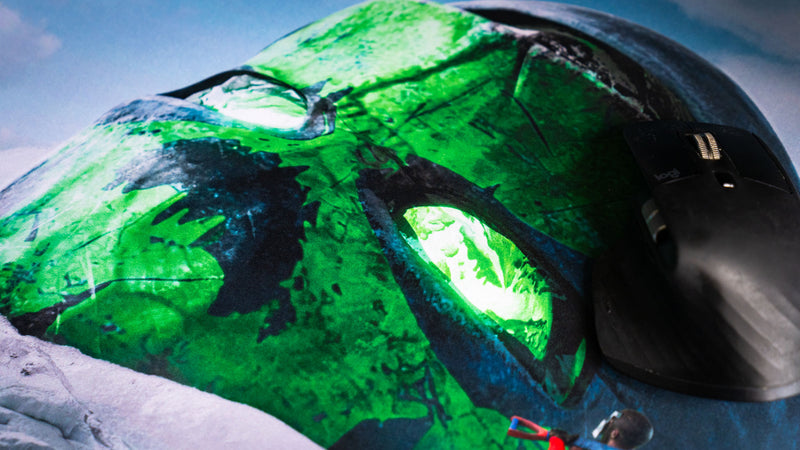 Close-up of the Oilrats Frozen Paradise deskmat featuring a weathered green glowing skull emerging from snow and ice, with a gaming mouse positioned over the artwork.