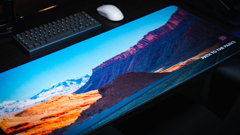 Angled close-up of the Path to the Parks America 250 deskmat featuring a bright Utah canyon road scene with red cliffs, snowy mountains, and bold landscape detail.