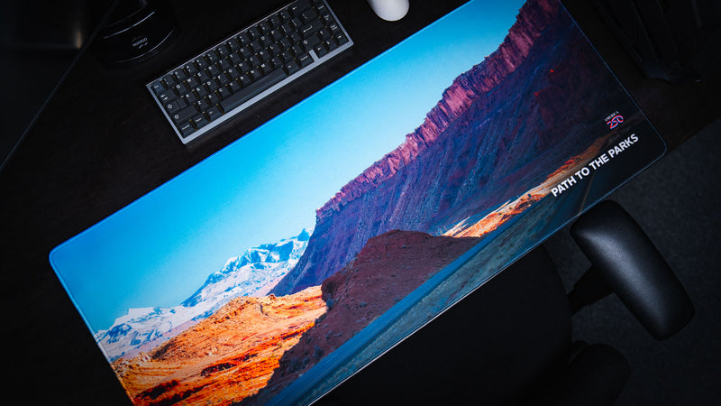 Diagonal setup view of the Path to the Parks America 250 deskmat featuring Utah red rock cliffs, snowy mountains, a desert road, and bright blue sky artwork.