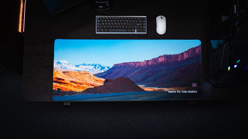 Overhead view of the Path to the Parks America 250 deskmat featuring a rural Utah road, red rock canyon cliffs, snowy mountains, and bright blue sky artwork.