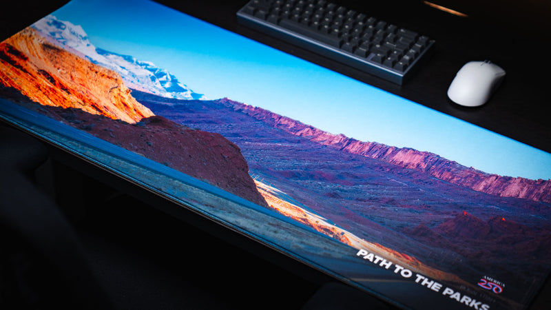 Angled view of the Path to the Parks America 250 deskmat featuring a rural Utah road, red rock cliffs, snowy mountains, and vivid desert colors.