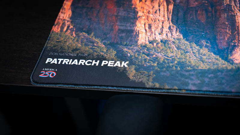 Close-up of the Patriarch Peak deskmat highlighting Zion National Park branding, the America 250 logo, premium stitched edges, and crisp HD print quality.
