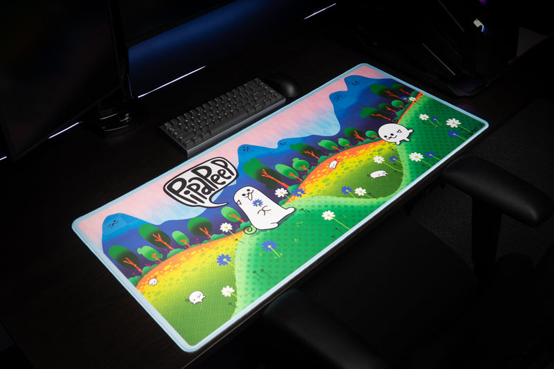 Angled view of the Pipaland deskmat on a gaming desk, showing the colorful hills and light-blue stitched edge.